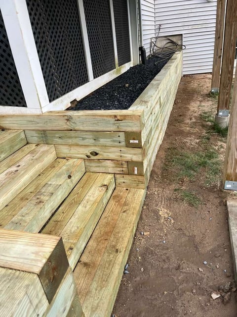 Wooden raised garden bed with black landscaping fabric and mulch against a white house foundation and siding