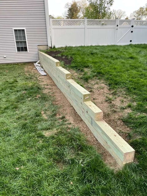 Wooden landscape edging installed along grass yard beside white vinyl fence and residential house