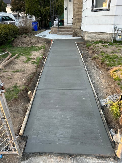 Newly poured concrete walkway leading to a house entrance, flanked by bare soil and landscaping on either side