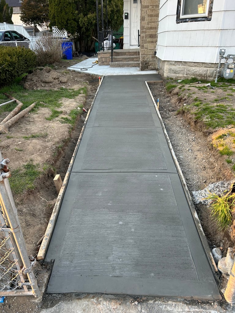 Newly poured concrete walkway leading to a house entrance, flanked by bare soil and landscaping on either side