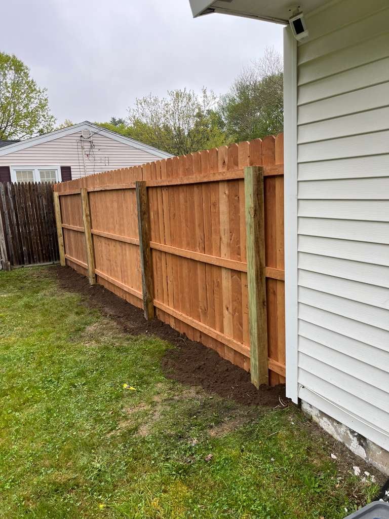 Wooden fence line in backyard next to white house siding with trees and residential home visible in background