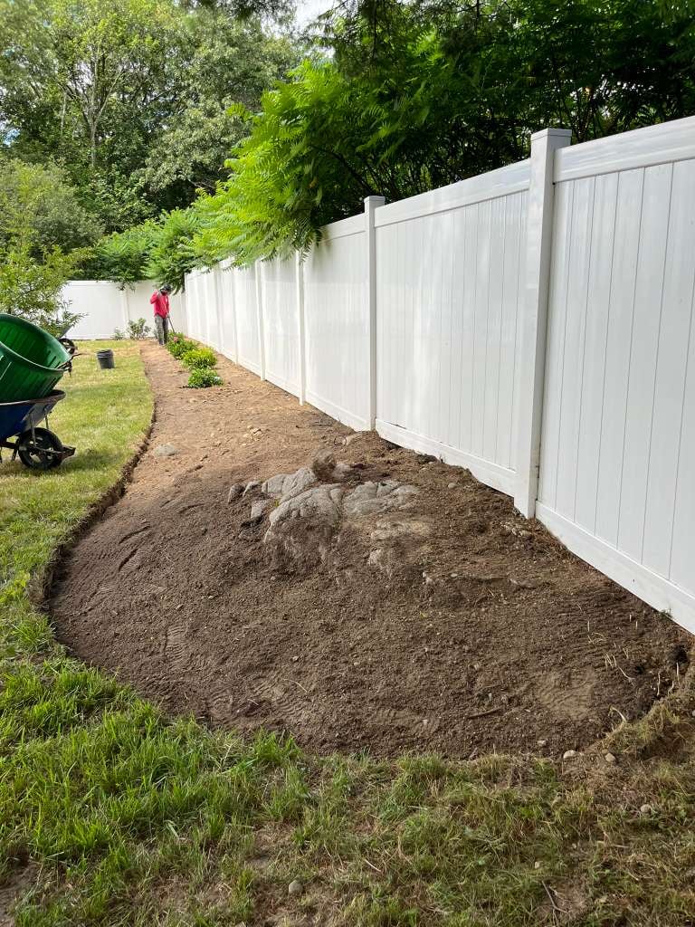 White vinyl fence along freshly dug dirt area in a residential backyard with green grass and trees