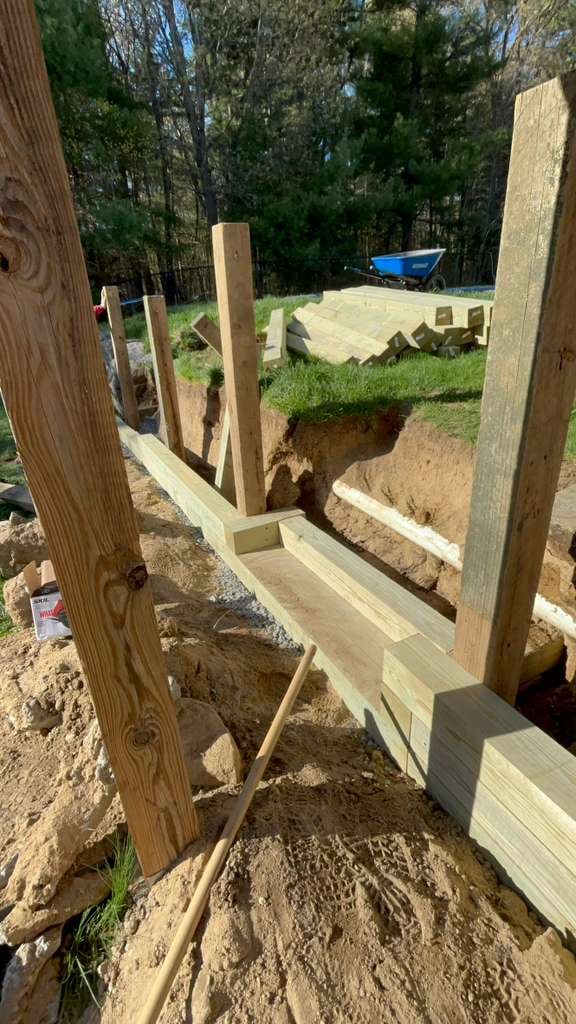 Construction of wooden framing structure in yard with excavated soil, trees, and wheelbarrow in background